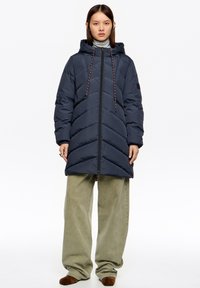 Kobieta ubrana w granatową pikowaną kurtkę z kapturem, jasnoszary golf, luźne oliwkowe spodnie i brązowe buty, stoi na tle białego tła.