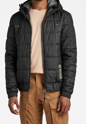 Homme portant une veste matelassée noire à capuche sur un t-shirt beige et un pantalon cargo marron avec poches zippées et à rabat.