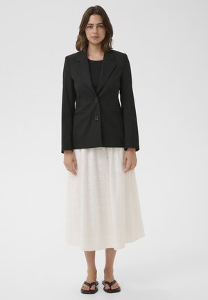 Vrouw draagt een zwarte blazer met twee knopen, een zwarte top, een witte midi-rok met patroon en zwarte open teen sandalen op een egale lichte achtergrond.