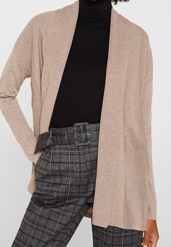 Beżowy kardigan o otwartym splocie z żebrowanymi rękawami, zestawiony z czarnym golfem i szarymi kraciastymi spodniami z paskiem i bocznymi kieszeniami.
