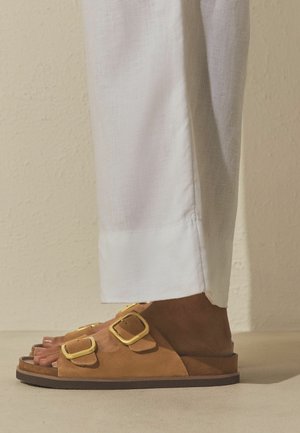 Nohy v hnědých semišových sandálech se dvěma zlatými přezkami, doplněné bílými lněnými kalhotami na pozadí béžové texturované stěny.