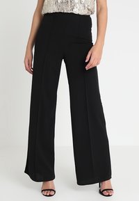 Svarta vida byxor i slät tyg. Har hög midja och pressade veck. Kombineras med en paljett topp och svarta sandaletter med remmar.