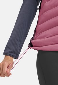 Giacca leggera isolante di colore viola con finitura ripstop, dotata di cordino regolabile all'orlo e abbinata a una maglietta a maniche lunghe di colore blu navy con texture.