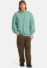 Mint green hoodie with front pocket and logo detail, paired with dark brown pants and black sneakers, topped with a brown knit beanie.