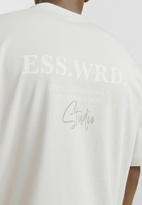 Lysbeige t-shirt med rund hals. Tekstprint på ryggen inkluderer "ESS.WRD" og "Studio" i kontrasterende hvid og grå.