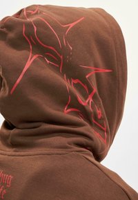 Felpa con cappuccio marrone caratterizzata da un grande design grafico rosso sulla schiena. Il tessuto sembra morbido e ha una texture liscia.
