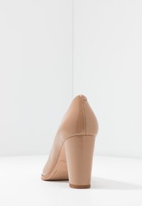 Chaussure à talons hauts en cuir verni beige avec un bout pointu et un talon épais. Texture lisse et design minimaliste.