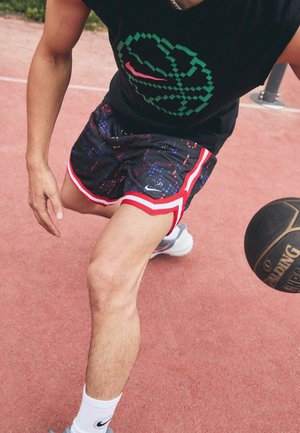 Młoda osoba w czarnej koszulce z nadrukiem i wzorzystych szortach dryblująca piłką do koszykówki Spalding na zewnętrznym boisku.