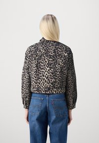 En person med långt blont hår bär en kort leopardmönstrad jacka och blåjeans, vänd bort från kameran mot en ljus bakgrund.