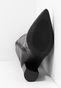 Czarny skórzany botek na obcasie ustawiony podeszwą do góry w białym rogu, ukazujący teksturowaną podeszwę i zamek błyskawiczny po boku.