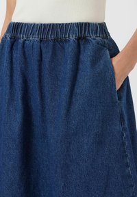 Jupe en denim avec une taille élastique, couleur bleu foncé et poches latérales. Le tissu a une texture douce et un léger éclat.