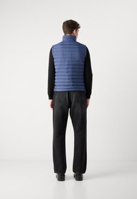 Chaleco acolchado azul con rayas horizontales, cuello alto y cierre de cremallera, usado sobre una camisa negra de manga larga y jeans negros.