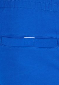 Modra tkanina z gladko teksturo ima vodoravno žep s šivanim robom. V žepu je vidna bela oznaka.