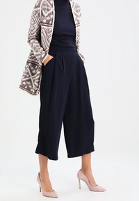 Culotte blu navy con pieghe, abbinate a un maglione testurizzato blu navy e a un cardigan aperto a fantasia. Tacchi a punta in tonalità neutre completano l'outfit.