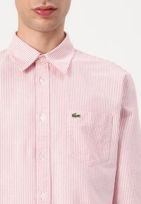Rødt og hvitt stripet skjorte med knappemerker og kragehals, med en lomme på venstre bryst og en grønn Lacoste-logo. Lett stoff.