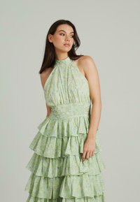 Zielona kwiatowa sukienka na ramiączkach z falbanami, teksturowany materiał i dopasowana talia. Posiada delikatne odcienie i subtelne wzory.