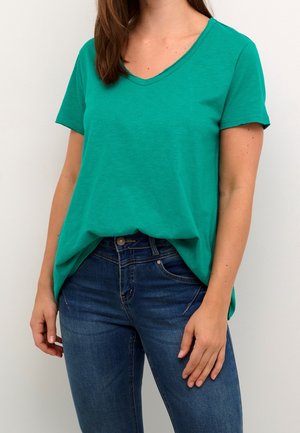 Femme portant un haut vert à col en V et manches courtes, légèrement rentré dans un jean en denim bleu sur un fond clair.