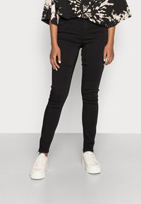 Calças skinny pretas com uma textura suave, cintura alta e design de cinco bolsos, combinadas com ténis brancos com detalhes verdes.