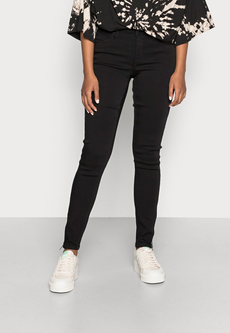 Calças skinny pretas com uma textura suave, cintura alta e design de cinco bolsos, combinadas com ténis brancos com detalhes verdes.