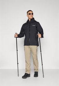 Black waterproof jacket with zip pockets, beige pants, and black hiking shoes. Accessories include trekking poles and sunglasses.