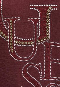 Il tessuto marrone scuro presenta lettere borchiate adornate con strass rossi, dorati e trasparenti, creando un effetto testurizzato e decorativo.