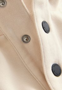 Gros plan d'un tissu beige avec des boutons pression noirs embossés "BAKER" et des boutons métalliques sur un vêtement.