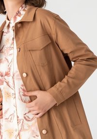 Giacca leggera marrone con chiusura a bottone e due taschini sul petto, indossata sopra una camicia a fantasia floreale in colori chiari.