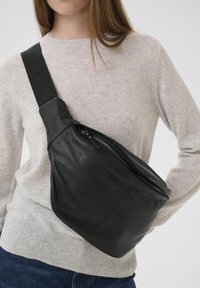 Una borsa a tracolla in pelle nera presenta una forma curva, una texture liscia e una tasca con zip. Indossata sopra un maglione grigio chiaro.