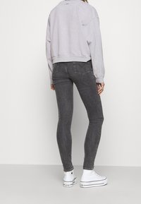 Persona vistiendo jeans ajustados grises, sudadera gris clara y zapatillas altas blancas de pie contra una pared blanca lisa.