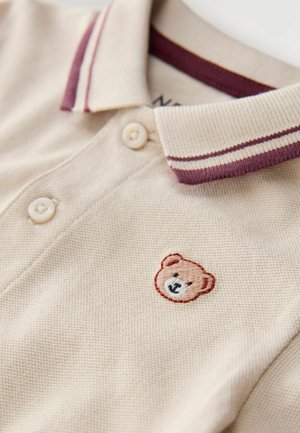 Polo color beige con tejido texturizado, con un cuello a rayas de color granate y un logo de oso bordado en el pecho. Incluye tres botones.