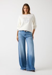 Maglione bianco con logo, abbinato a jeans chiari a vita alta e a gamba ampia. Tessuto morbido, vestibilità rilassata, caratterizzato da un sottile gradiente.