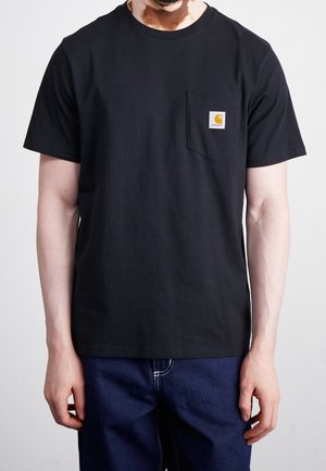 Homme atteint de vitiligo portant un t-shirt noir Carhartt à manches courtes avec poche poitrine et jean bleu foncé sur un fond uni.