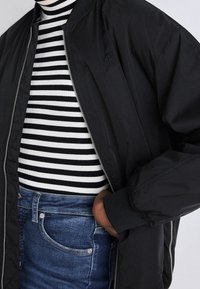 Persoon in een zwarte zip-up jas, een zwart-wit gestreepte coltrui en blauwe spijkerbroek met een hand in de jaszak gestoken.
