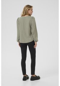 Groene blouse met stippen, pofmouwen en een ronde halslijn, gecombineerd met zwarte slim-fit broek en zwarte platform schoenen.