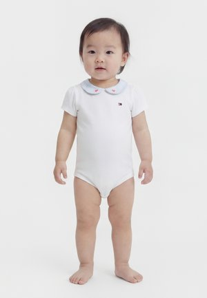 Kleuter staat op blote voeten en draagt een wit rompertje met korte mouwen, een blauw gestreepte Peter Pan-kraag en kleine hartjesdetails.