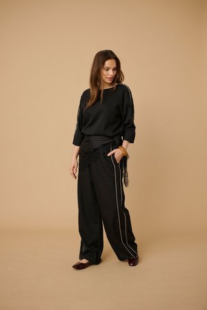 Frau trägt schwarze locker sitzende Hose mit weißen Seitenstreifen, ein passendes schwarzes Oberteil und bordeauxrote flache Schuhe, steht vor beigem Hintergrund.