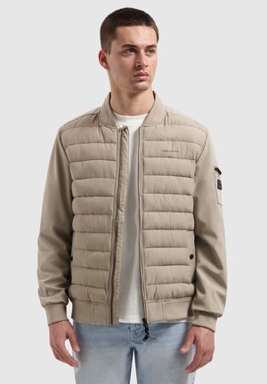 Junger Mann blickt nach rechts, trägt eine beige gesteppte Bomberjacke über einem weißen Hemd und hellblauen Jeans, steht vor einem schlichten Hintergrund.