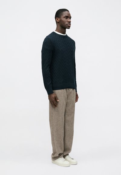 Pull en maille texturée bleu marine avec un col rond côtelé, associé à un pantalon large beige et des baskets blanches.