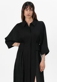 Vestito camicia nero con colletto, apertura frontale con bottoni e maniche corte. Presenta una vita con cintura, tasche laterali e uno spacco frontale per un tocco in più.