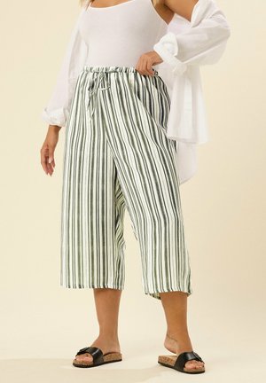 Kobieta ubrana w krótkie, szerokie spodnie w paski, biały top na ramiączkach i białą koszulę, do tego czarne klapki na beżowym tle.