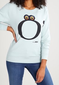 Vrouw in een lichtblauwe sweatshirt met een groot zwart abstract kikkerontwerp en kleurrijke ogen, gecombineerd met blauwe spijkerbroek.