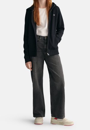 Persoon draagt een zwarte hoodie met rits, wit shirt, losse donkergrijze jeans en witte sneakers met rode accenten, staand tegen een effen achtergrond.