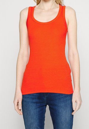 Kobieta ubrana w jasnoczerwoną, bezrękawnikową, prążkowaną koszulkę w połączeniu z ciemnoniebieskimi dżinsami na jednolitym tle.