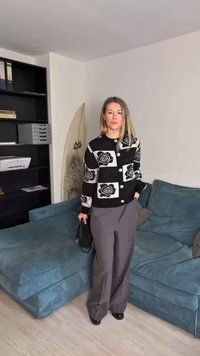Črno-bela kardigan z cvetličnimi motivi in velikimi gumbi, v kombinaciji z sivimi širokimi hlačami. Črna torbica, modra zofe v ozadju.
