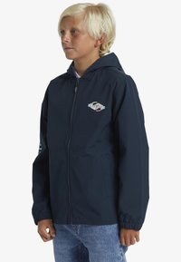 Mørkeblå zip-up jakke med hætte, har elastiske manchetter og et logo på brystet. Konstrueret af letvægts, vandafvisende materiale.