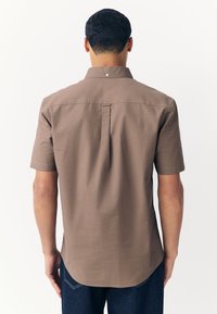 Camisa marrón de manga corta con corte recto, pinza en la parte trasera y detalle sutil en el cuello. La tela parece suave y de estilo casual.