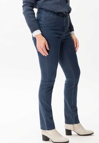 Jean skinny en denim bleu foncé avec une taille mi-haute, poches avant et une texture lisse, associés à des bottines crème.