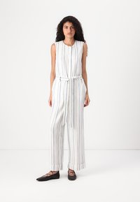 Stripet linne jumpsuit med knapper foran, ermeløs design og knyttet liv. Har vertikale blå og hvite striper. Flate svarte sko.