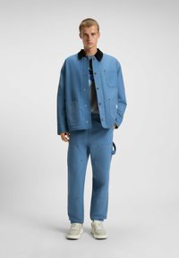 Blauwe katoenen jas met donkere kraag, knoopsluiting en twee voorzakken; gecombineerd met bijpassende blauwe broek met zichtbare stiksels en zakken.