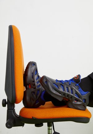 Des pieds portant des baskets bleu et noir reposant sur le siège d'une chaise de bureau orange sur fond blanc.